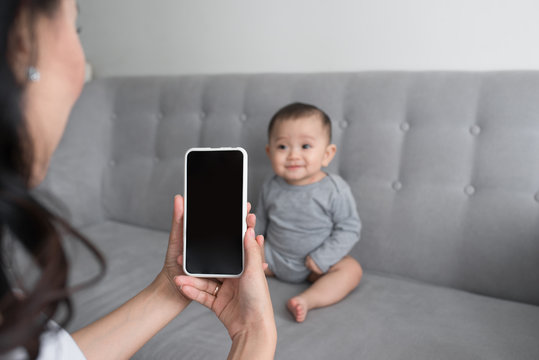 Happy mother taking a picture of her baby girl on sofa with smart phone