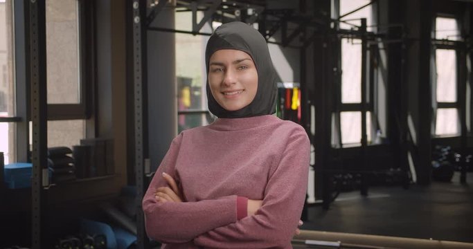 Closeup portrait of young attractive athletic muslim female looking at camera with her arms crossed over chest smiling cheerfully in gym indoors