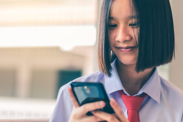 Smiling female Asian high school student in white uniform is enjoying social media on her smartphone.