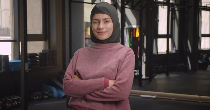 Closeup portrait of young attractive athletic muslim female looking at camera with her arms crossed over chest smiling cheerfully standing in gym indoors