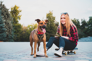 Teenage skater girl having fun with her dog in the city. Mixed breed dog stands next to young woman...