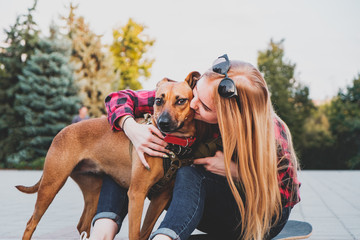 Teenage skater girl kissing her dog in urban environment. Mixed breed dog stands next to young...