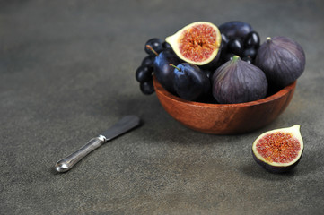 Figs, plums and grapes in a wooden bowl. Half a fig on the table. The concept of purple fruits. Dark background. Close-up. Free space for text.