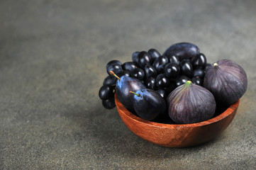 Figs, plums and grapes in a wooden bowl. The concept of purple fruits. Dark background. Close-up. 