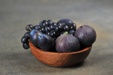 Figs, plums and grapes in a wooden bowl. The concept of purple fruits. Dark background. Close-up. Free space for text.