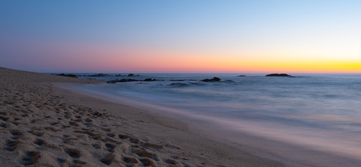 Long exposure shot over beach at dusk with milky ocean and smooth blue orange gradient sky. Dark rocks in water.