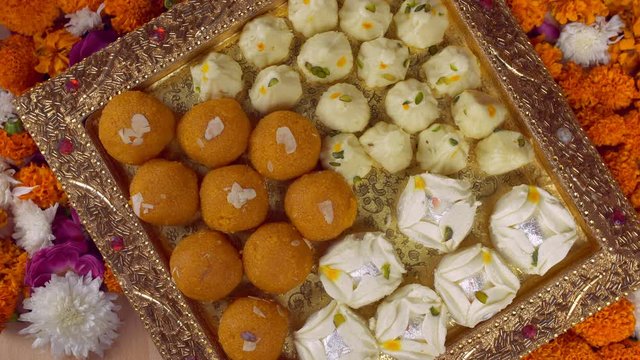 Top view of a decorated plate with Indian sweets on the occasion of Diwali - the festival of India. Silver rotating tray with Modak  Chamcham  Bundi Laddu/Motichoor on a decorated floral platform f...