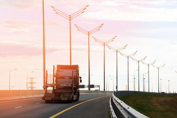Highway view against evening sky, row of lampposts, empty car transporter truck driving away in sun glare