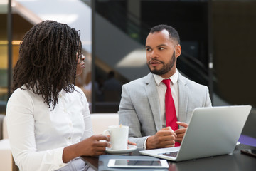 Diverse couple of colleagues watching and discussing content on laptop. Business man and woman drinking coffee, using computer and talking. Digital communication concept