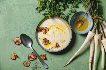 Parsnip cream soup in ceramic bowl with sun dried pears, butter sauce, bundle of fresh parsnip and herbs over green texture background. Flat lay, space - obrazy, fototapety, plakaty