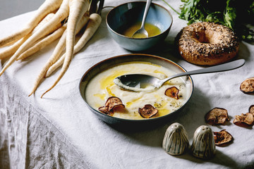 Parsnip cream soup in ceramic bowl with butter sauce, sun dried pears, bundle of fresh parsnip, bagel bread and herbs on kitchen table with white tablecloth