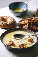 Parsnip or cauliflower cream soup in ceramic bowl with butter sauce, sun dried pears, bagel bread and herbs on kitchen table with white tablecloth