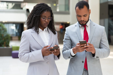 Serious multiethnic business colleagues consulting internet on mobile phones. Business man and woman standing in office hallway, using smartphones, texting message. Digital communication concept