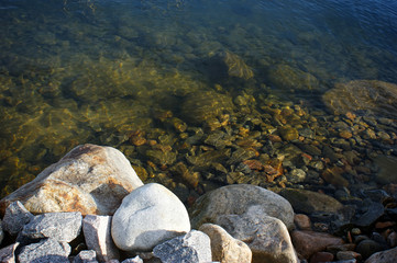 Photo through the water. Photo of the bottom of the lake with granite stones. On the shore lies a mountain of stones.