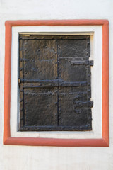 Ancient metal shutters on a window with forged details. The surface is painted black. Window framed with red ledge on white wall