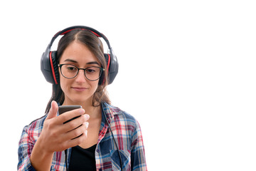 Caucasian young girl with plaid shirt and glasses, listening to music with big headphones and a mobile phone or other type of electronic device on white background isolated