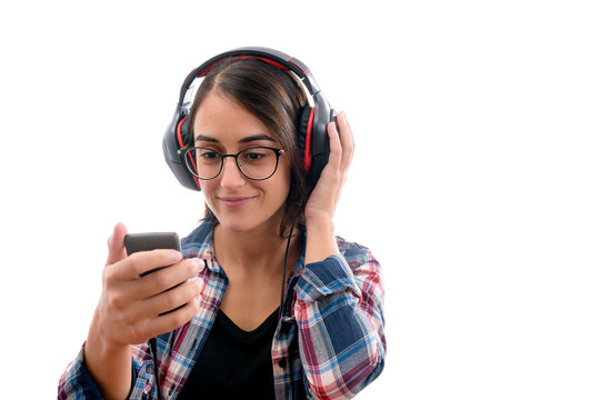 Caucasian young girl with plaid shirt and glasses, listening to music with big headphones and a mobile phone or other type of electronic device on white background isolated