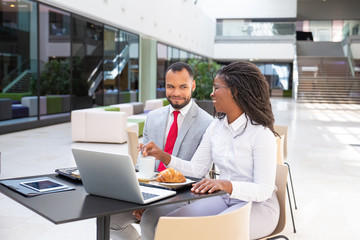 Happy business colleagues having breakfast in cafe. Business man and woman sitting at laptop outside and talking. Business breakfast concept