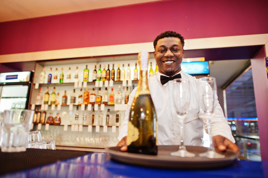 African american bartender at bar holding champagne with glasses on tray.