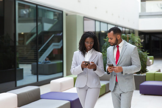 Focused excited business colleagues using mobile phones while discussing work issues. Business man and woman walking through office hallway and holding smartphones. Wireless communication concept