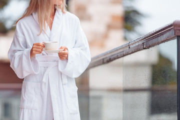 Charming healthy young blonde woman with a cup of coffee in a bathrobe walks barefoot on her large terrace enjoying the summer nature. Concept o vacation in own big country house or hotel - obrazy, fototapety, plakaty
