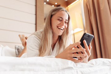 Fototapeta na wymiar Bottom view of laughing cheerful beautiful young woman watching private messages in social networks and enjoying lying on bed in spa hotel in white bathrobe. Concept of high speed internet and gadgets