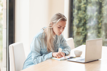 Beautiful attentive girl student preparing for exams by writing notes in a notebook using a laptop and high-speed Internet while sitting at the kitchen table in the morning