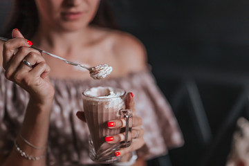 young woman sitting in cafe holding spoon ang glass with coffee or milkshake with whipped cream