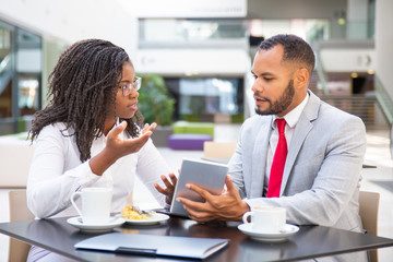 Businessman showing project presentation to female colleague. Diverse business man and woman sitting in cafe, using tablet together and talking. Digital communication concept