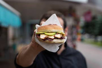 Young man is holding a juicy chicken burger in front of his face during street photoshoot