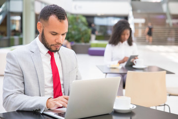 Fototapeta na wymiar Focused serious businessman using laptop in co-working space. Young African American woman using digital device in background. Workspace concept