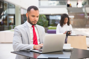 Focused male business leader working on laptop in coffee shop. Young African American woman using digital device in background. Internet concept