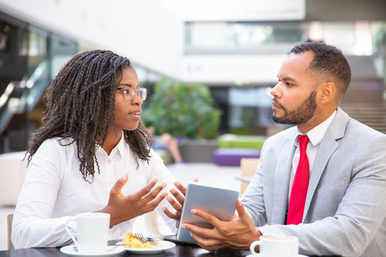 Excited business colleagues discussing tablet app. Diverse business man and woman sitting in cafe, using tablet together and talking. Digital communication concept