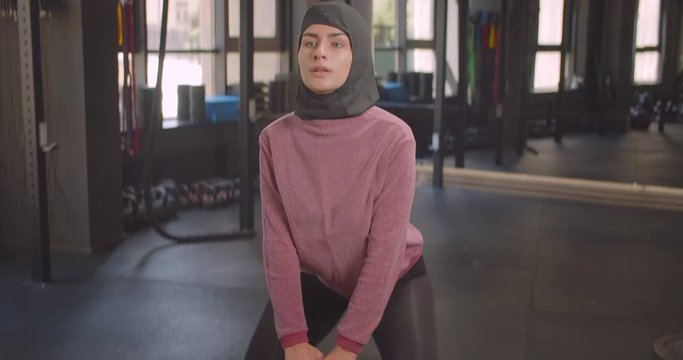 Closeup portrait of young determined athletic muslim female making squats with dumbbell in gym indoors