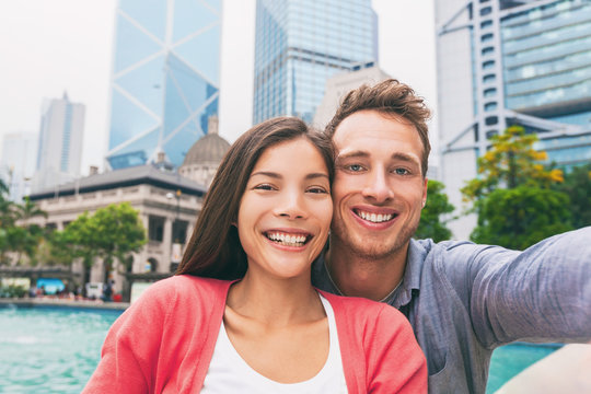 Selfie travel photo with phone of young adults tourists in love smiling at camera. Interracial couple in Hong Kong city, urban living. Asian multiracial chinese woman, Caucasian man in their 20s.