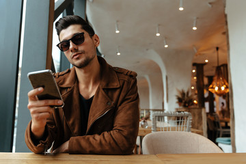 City style. Technology. Cafe. Handsome young man in casual clothes and sunglasses is using a smartphone while sitting in cafe