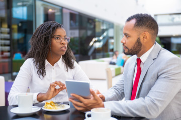 Diverse business team watching content on tablet and discussing project. Business man and woman sitting in cafe, using tablet together and talking. Corporate brainstorming concept