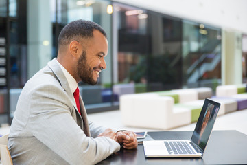 Fototapeta premium Smiling young businessman chatting via laptop. Cheerful African American businessman using laptop computer and having video chat with colleague at lunchtime. Communication concept