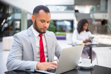 Confident serious executive working during coffee break. Business man sitting at table and using laptop. Hardworking business leader concept