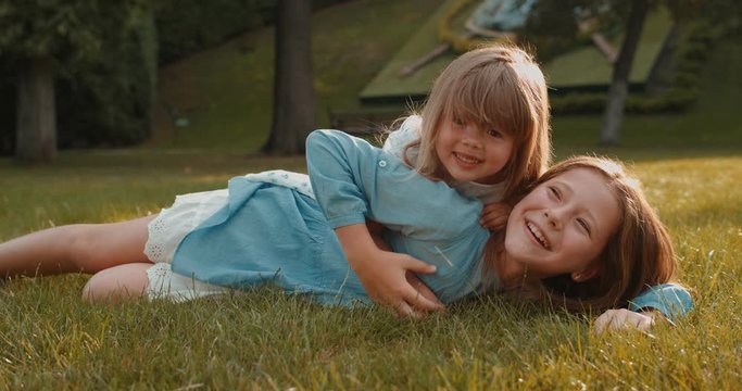 Portrait shot of the cute and pretty Caucasian teen girl lying on the green grass in the park and her small younger sister lying on her back, they both smiling to the camera and having fun.