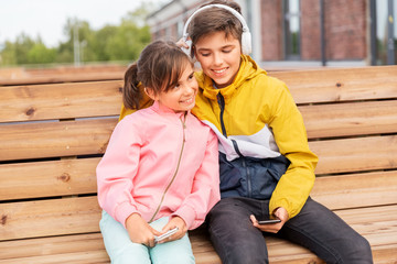 childhood, technology and people concept - happy children or brother and sister with headphones and smartphones sitting on wooden street bench outdoors and listening to music