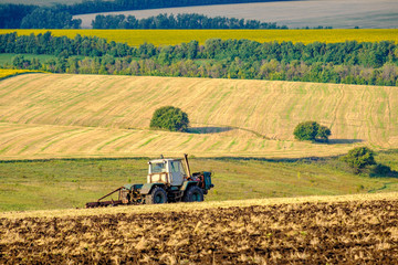 An agricultural tractor plows a field with a plow after harvested wheat. In the background are...