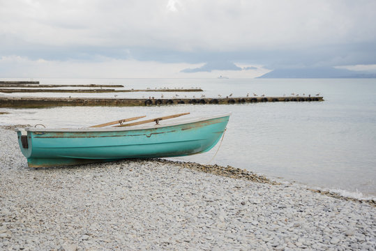 side view of small wooden fishing azure boat on pebble coast black sea beach in bad weather on dramatic sea and sky background, horizontal stock photo image