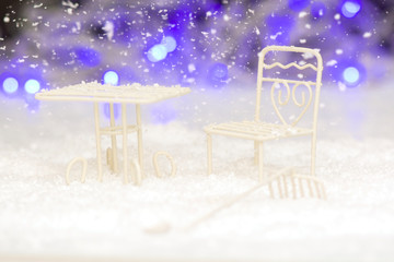 Christmas scene of white decorative table and chair, buried in decorative snow on the background of blurred neon lights.