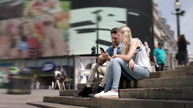 Urban lifestyle. Happy young couple sitting in Piccadilly Circus in London. Beautiful young couple dating smiling and watching messages and pictures on a smartphone in UK. Blond woman, handsome man.