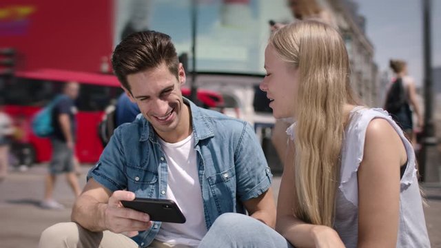 Portrait of happy young couple sitting in Piccadilly Circus in London. Beautiful young couple dating smiling and watching messages and pictures on a smartphone in UK. Blond woman, handsome man.