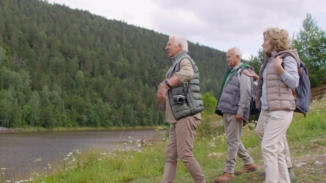 Tracking shot of group of elderly tourists with backpacks walking towards river and enjoying view on their hike. Senior man with grey hair and mustache taking picture on digital camera
