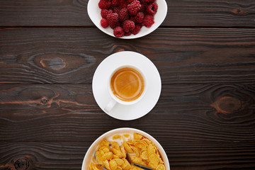 top view of bowl with cornflakes and milk near cup of coffee and plate of raspberry on wooden surface