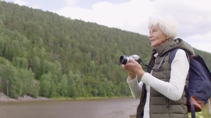 Tracking shot of cheerful senior woman with backpack standing by river and taking picture on digital camera during hike on chilly summer day