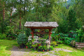 Naklejka premium Old retro stone well with a wooden roof with lots of flower pots and small trees in a botanical garden on a summer day. Green fresh plants on the nature.